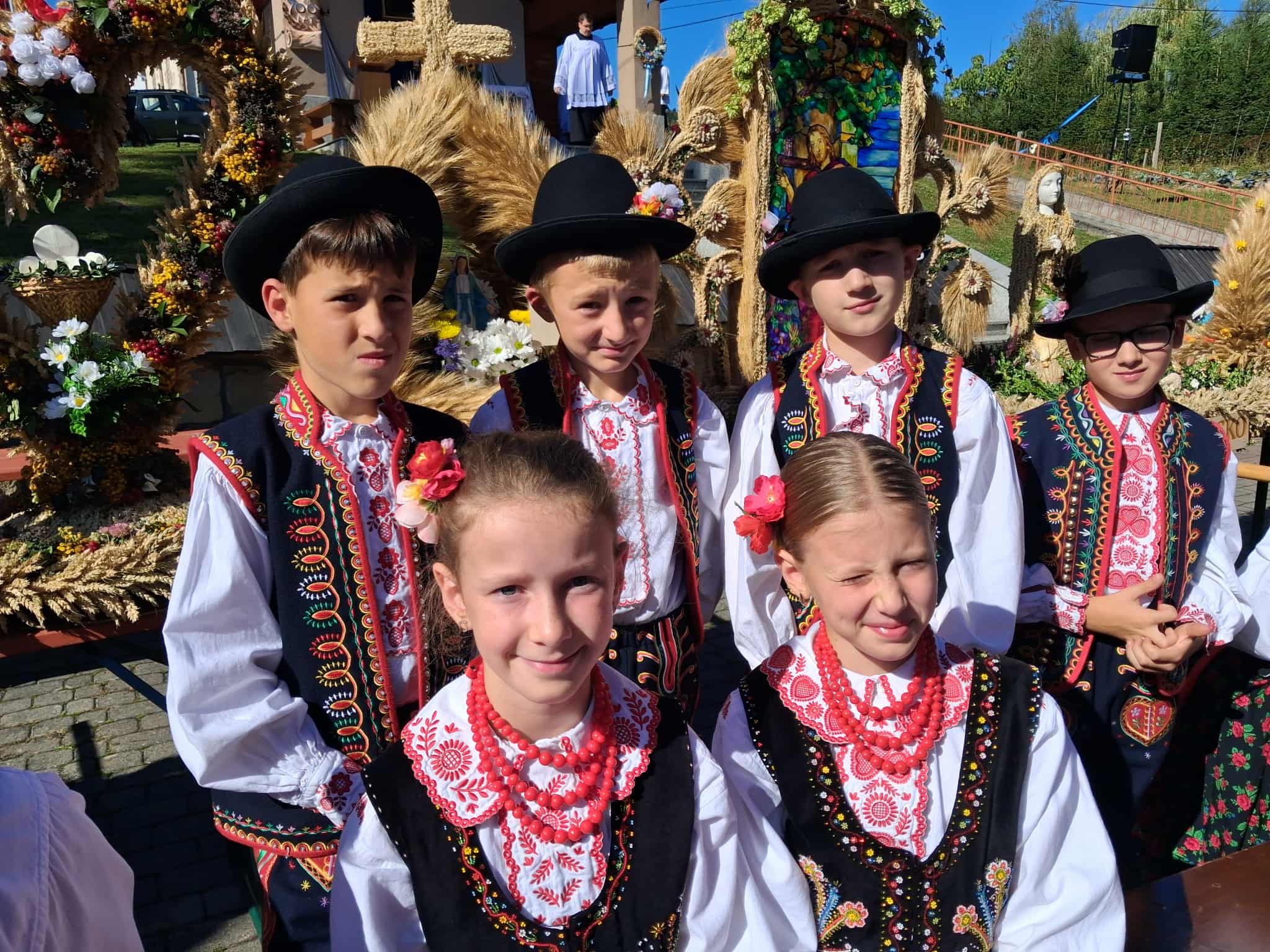 Zdjęcie zbiorowe uczniów - czterech chłopców i 2 dziewczynek w strojach regionalnych. W tle wieńce dożynkowe