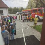 Na zdjęciu: osoby na parkingu, wóż strażacki i strażacy, rozciągnięty wąż