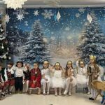 Zdjęcie panoramiczne - pani dyrektor wita wszystkich przybyłych, dzieci w strojach, w tle dekoracja: ozdobne płótno przedstawiające choinki zimą, po bokach niebieskie płótno, gwiazdki, choinka, szopka ze żłóbkiem