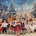 Dzieci w strojach siedzą na ławeczce i machają