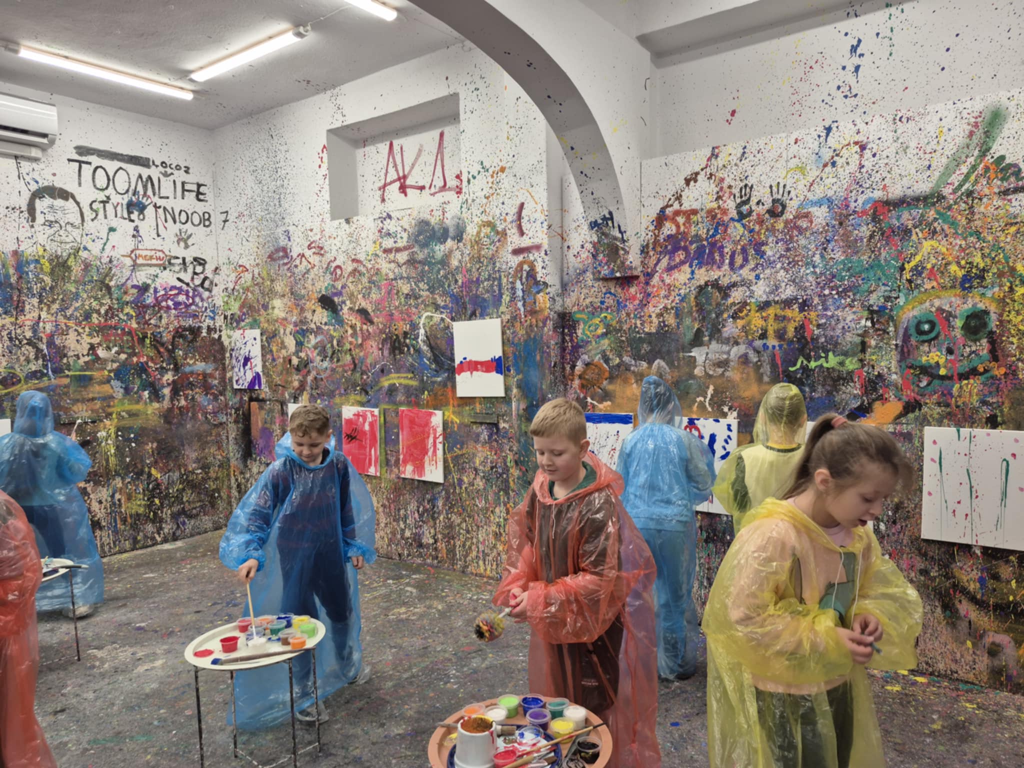 Uczniowie w Pokoju Ekspresji - ubrani w peleryny, na ścianach rozwieszona płótna na obrazy, które uczniowie malują, ściany pochlapane i pomalowane farbami