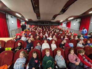 Sala widowiskowa wypełniona dziećmi siedzącymi na czerwonych fotelach, widok od strony sceny w głąb widowni. Dzieci patrzą w stronę aparatu, część się uśmiecha, a po bokach sali stoją dorośli opiekunowie. Wnętrze ma czerwone zasłony i jasny sufit.