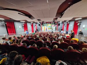 Widok z tyłu sali teatralnej wypełnionej dziećmi siedzącymi na czerwonych fotelach. Na scenie widać kolorową scenografię i osoby przygotowujące się do przedstawienia, a po bokach sali stoją dorośli opiekunowie.