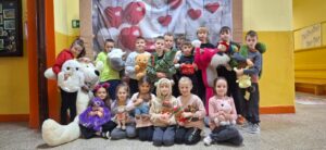 Grupa małych dzieci stoi i siedzi razem do wspólnego zdjęcia w sali szkolnej. Każde trzyma pluszową zabawkę, a z przodu leży bardzo duży biały miś. W tle widać dekorację z czerwonych i białych serc.