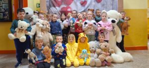 Grupa małych dzieci stoi i siedzi razem do wspólnego zdjęcia w sali szkolnej. Każde trzyma pluszową zabawkę, a z przodu leży bardzo duży biały miś. W tle widać dekorację z czerwonych i białych serc.