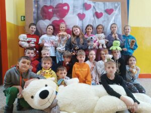 Grupa małych dzieci stoi i siedzi razem do wspólnego zdjęcia w sali szkolnej. Każde trzyma pluszową zabawkę, a z przodu leży bardzo duży biały miś. W tle widać dekorację z czerwonych i białych serc.