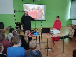 Grupa dzieci siedzi na ławkach w sali gimnastycznej i ogląda prezentację na dużym ekranie. Przed nimi stoi mężczyzna, który prowadzi spotkanie. 