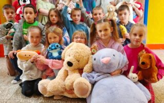 Grupa małych dzieci stoi i siedzi razem do wspólnego zdjęcia w sali szkolnej. Każde trzyma pluszową zabawkę, a z przodu leży bardzo duży biały miś. W tle widać dekorację z czerwonych i białych serc.