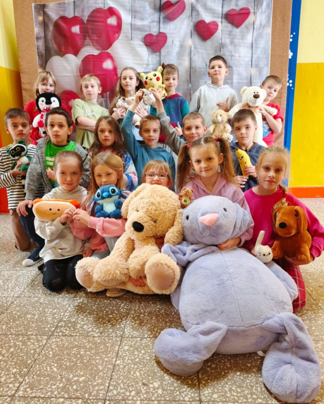 Grupa małych dzieci stoi i siedzi razem do wspólnego zdjęcia w sali szkolnej. Każde trzyma pluszową zabawkę, a z przodu leży bardzo duży biały miś. W tle widać dekorację z czerwonych i białych serc.