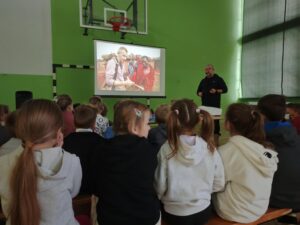 Grupa dzieci siedzi na ławkach w sali gimnastycznej i ogląda prezentację na dużym ekranie. Przed nimi stoi mężczyzna, który prowadzi spotkanie. 