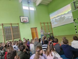Na zdjęciach widać dzieci siedzące na materacach w sali gimnastycznej. Słuchają z zaciekawieniem Pana podróżnika który opowiada o swoich wyprawach w góry. Pokazuje zdjęcia oraz filmy z wypraw.