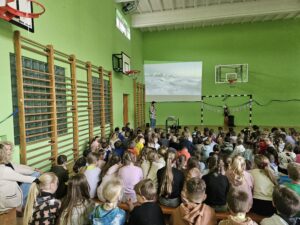 Na zdjęciach widać dzieci siedzące na materacach w sali gimnastycznej. Słuchają z zaciekawieniem Pana podróżnika który opowiada o swoich wyprawach w góry. Pokazuje zdjęcia oraz filmy z wypraw.