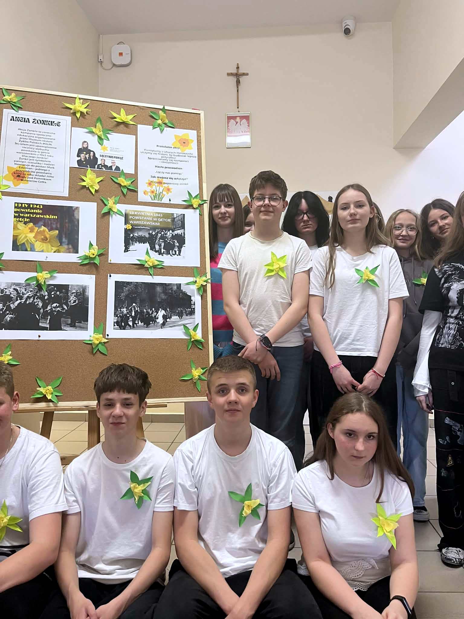 Uczniowie kl. 7 i 8 obok wystawki z żonkilami zrobionymi z papieru przypiętymi do białych podkoszulek